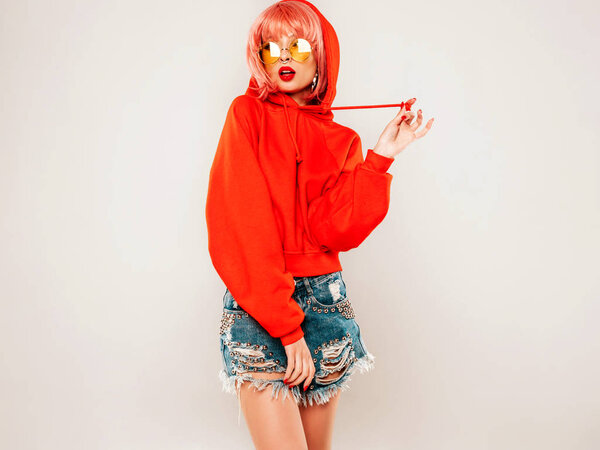 Young beautiful hipster bad girl in trendy red summer hoodie and earring in her nose.Sexy carefree woman in pink wig posing in studio on gray background.Hot model in sunglasses