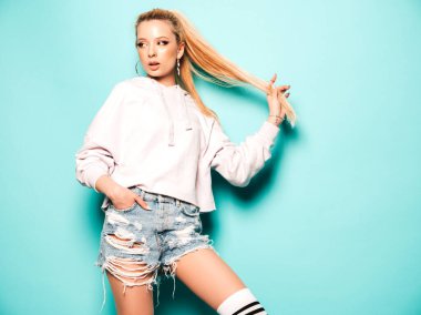 Yeni moda kot şort ve kapüşonlu kıyafetler giyen güzel, gülümseyen hippi kız portresi. Stüdyoda mavi duvarın yanında poz veren seksi, kaygısız kadın. Pozitif model eğleniyor. 