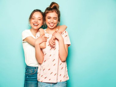 İki güzel sarışın, gülümseyen yenilikçi kız, son moda hippi kot pantolon giyiyorlar. Mavi duvarın yanında poz veren kaygısız seksi kadınlar. Moda ve pozitif modeller eğleniyor. Dillerini gösteriyorlar.