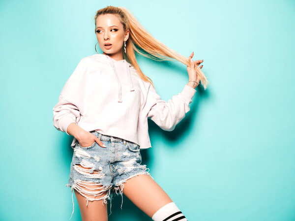 Portrait of young beautiful smiling hipster girl in trendy summer jeans shorts and hoodie clothes. Sexy carefree woman in gaiters posing near blue wall in studio. Positive model having fun 