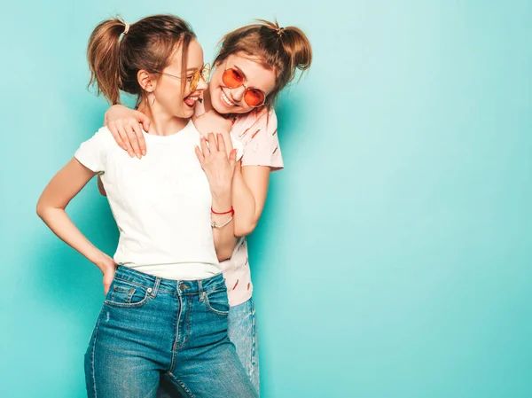 流行の夏のヒップスタージーンズの服で2人の若い美しいブロンドの笑顔ヒップスター女の子 青い壁の近くにポーズをとるセクシーな屈託のない女性 彼らは異言を示しています — ストック写真