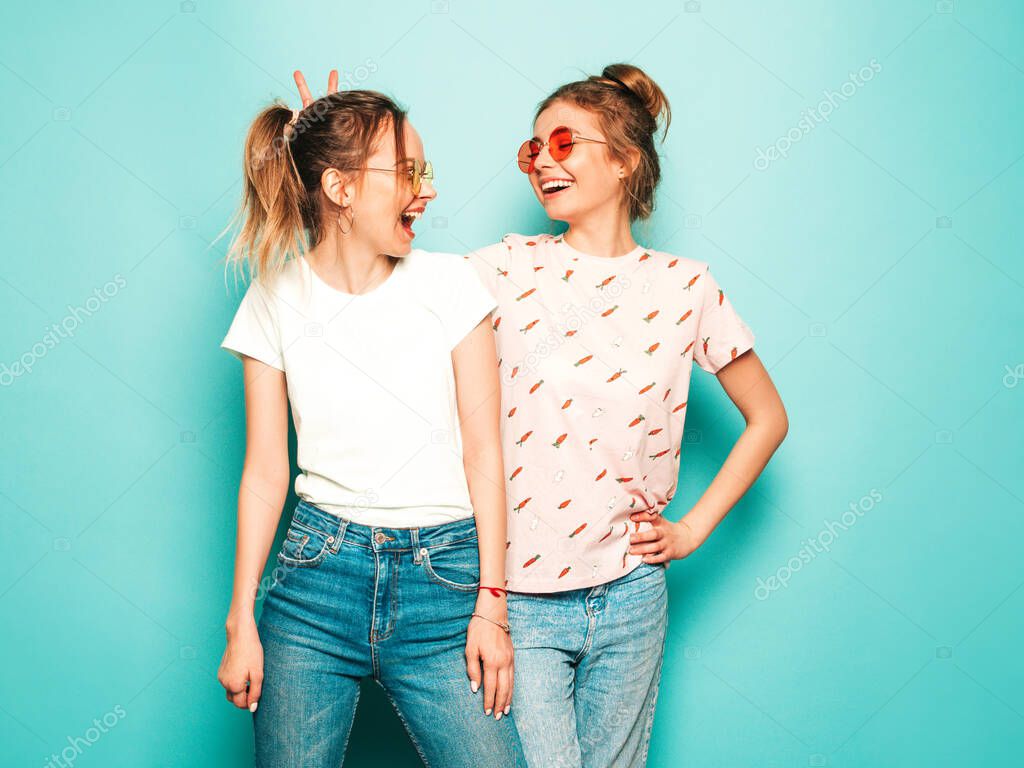 Two young beautiful blond smiling hipster girls in trendy summer hipster jeans clothes. Sexy carefree women posing near blue wall. Trendy and positive models having fun