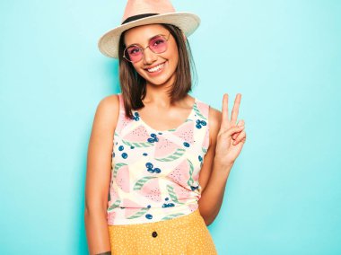 Şapkalı genç, güzel bir kadın. Gündelik beyaz tişörtlü, sarı etekli, güneş gözlüklü, modaya uygun bir kız. Pozitif kadın yüz duyguları gösterir. Barış işareti gösterir