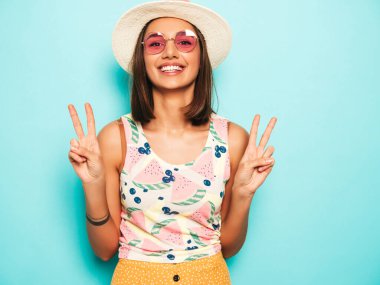 Şapkalı genç, güzel bir kadın. Gündelik beyaz tişörtlü, sarı etekli, güneş gözlüklü, modaya uygun bir kız. Pozitif kadın yüz duyguları gösterir. Barış işareti gösterir