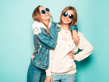 Son moda kapüşonlu ve kot ceketli gülümseyen iki güzel esmer kız. Mavi duvarın yanında poz veren kaygısız seksi kadınlar. Moda ve pozitif modeller güneş gözlükleriyle eğleniyor.