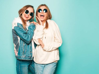 Son moda kapüşonlu ve kot ceketli gülümseyen iki güzel esmer kız. Mavi duvarın yanında poz veren kaygısız seksi kadınlar. Moda ve pozitif modeller güneş gözlükleriyle eğleniyor.