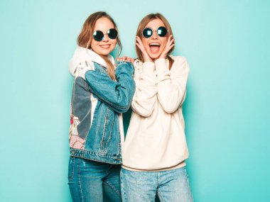 Son moda kapüşonlu ve kot ceketli gülümseyen iki güzel esmer kız. Mavi duvarın yanında poz veren kaygısız seksi kadınlar. Moda ve pozitif modeller güneş gözlükleriyle eğleniyor.