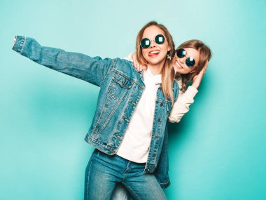 Son moda kapüşonlu ve kot ceketli gülümseyen iki güzel esmer kız. Mavi duvarın yanında poz veren kaygısız seksi kadınlar. Moda ve pozitif modeller güneş gözlüklü dili gösteriyor