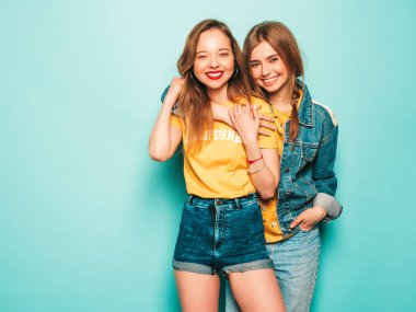 Son moda sarı tişörtlü ve kot ceketli iki güzel gülümseyen hippi kız. Mavi duvarın yanında poz veren kaygısız seksi kadınlar. Moda ve pozitif modeller eğleniyor.