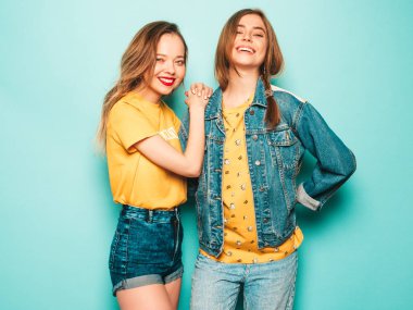 Son moda sarı tişörtlü ve kot ceketli iki güzel gülümseyen hippi kız. Mavi duvarın yanında poz veren kaygısız seksi kadınlar. Moda ve pozitif modeller eğleniyor.