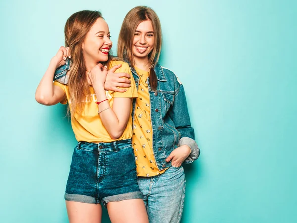 流行の夏の黄色のTシャツとジーンズのジャケットで2人の若い美しい笑顔のヒップスター女の子 青い壁の近くにポーズをとるセクシーな屈託のない女性 流行と肯定的なモデルは舌を示しています — ストック写真