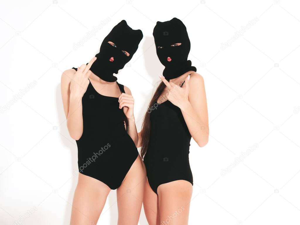 Two beautiful sexy women in black swimwear bathing suit. Models wearing bandit balaclava mask.Hot girls posing near white wall in studio.Seductive female in nice lingerie.Crime and violence