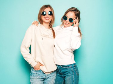Son moda yaz kapüşonlu giysileri içinde gülümseyen iki güzel sarışın kız. Mavi duvarın yanında poz veren kaygısız seksi kadınlar. Moda ve pozitif modeller güneş gözlükleriyle eğleniyor.