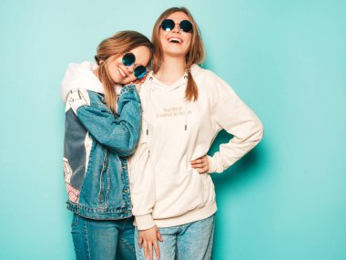 Son moda kapüşonlu ve kot ceketli gülümseyen iki güzel esmer kız. Mavi duvarın yanında poz veren kaygısız seksi kadınlar. Moda ve pozitif modeller güneş gözlükleriyle eğleniyor.