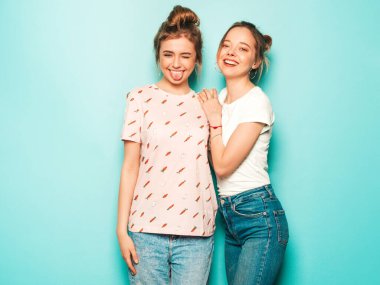 İki güzel sarışın, gülümseyen yenilikçi kız, son moda hippi kot pantolon giyiyorlar. Mavi duvarın yanında poz veren kaygısız seksi kadınlar. Moda ve pozitif modeller eğleniyor. Dillerini gösteriyorlar.