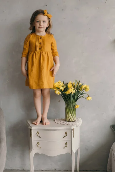 little girl in a yellow dress with a beautiful bow stands on a baroque nightstand near a bouquet of  narcissus flowers