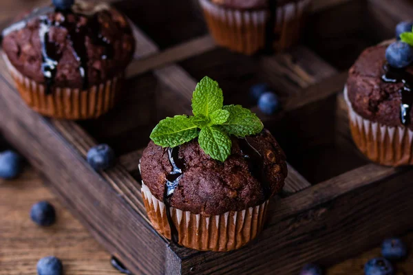 Chocolate muffins with chocolate syrup, blueberries and mint in a wooden background