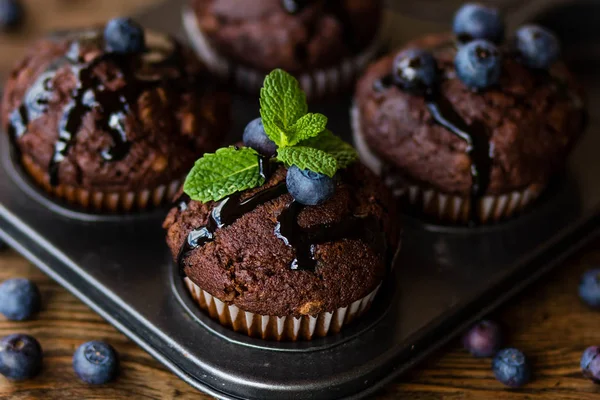 Chocolate muffins with chocolate syrup, blueberries and mint in a wooden background — Stock Photo, Image