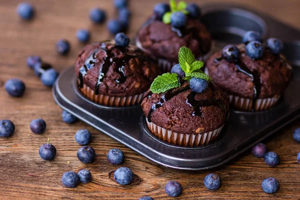 Muffins de chocolate com xarope de chocolate, mirtilos e hortelã em um fundo de madeira — Fotografia de Stock