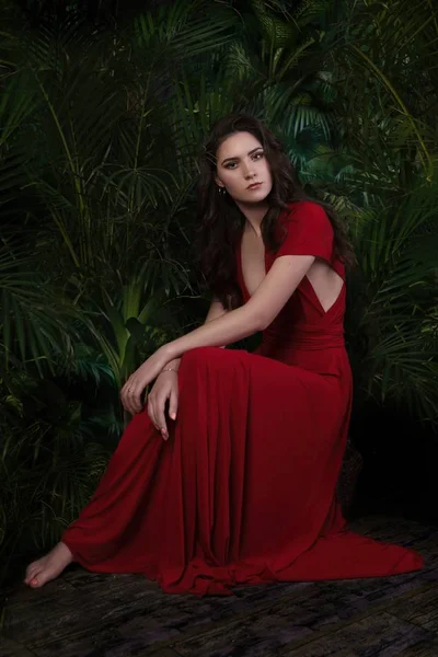 Dívka v červených šatech sedí v džungli mezi listy — Stock fotografie