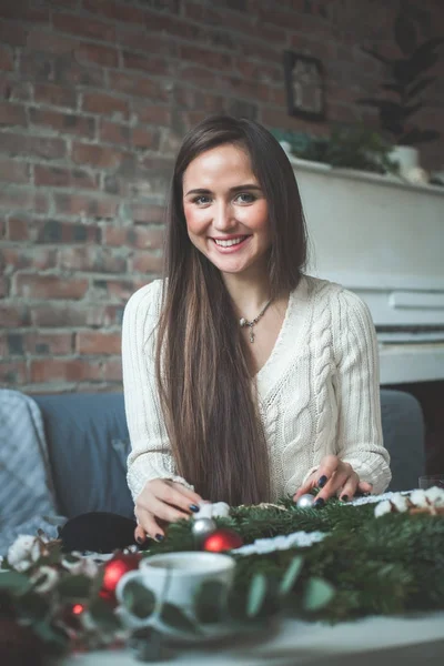 Mujer joven sonriendo y haciendo guirnalda de Navidad en casa —  Fotos de Stock