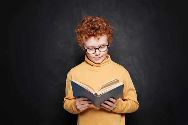 Porträt eines kleinen Jungen mit Brille, der ein Buch gegen die Tafel liest — Stockfoto