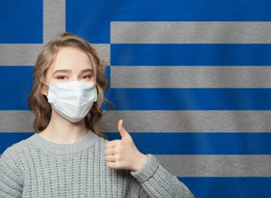 Yüz maskeli mutlu kadın, ulusal Yunan bayrağı arka planında başparmağını kaldırıyor. Grip salgını ve virüs koruma kavramı