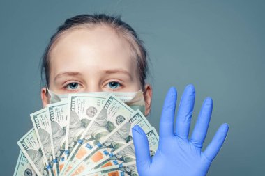 Tıbbi maskeli kız elinde para, Amerikan doları ve mavi arka planda durdurma hareketi gösteriyor.