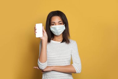 Tıbbi maske takan siyah bir kadın elinde parlak sarı arka planda boş ekranlı akıllı bir telefon tutuyor.