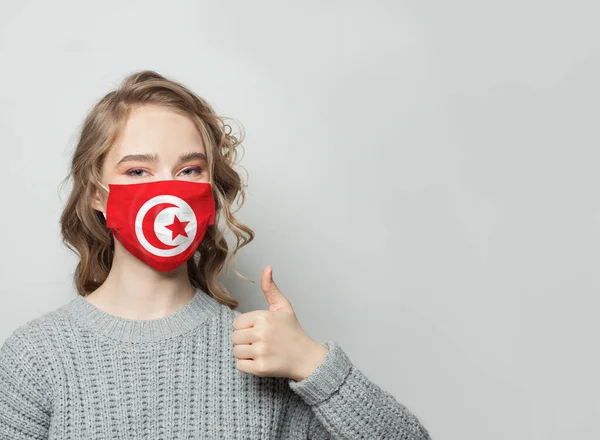 チュニジアの国旗を背景に親指を立てるフェイスマスクの幸せな女性 インフルエンザの流行とウイルス保護の概念 — ストック写真