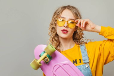 Güneş gözlüklü genç bir kadın beyaz arka planda kaykay tutuyor.