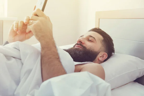 Szakállas férfi feküdt az ágyban reggel telefon — Stock Fotó