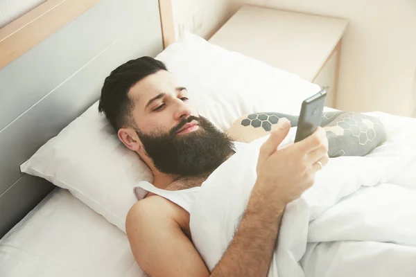 Bärtiger Mann liegt mit Handy im Morgenbett — Stockfoto
