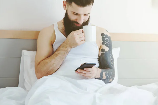 Bärtiger Mann liegt mit Morgenkaffee und Handy per App im Bett — Stockfoto