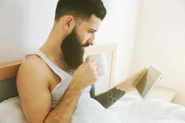 Bärtiger Mann im Morgenbett liegend mit Kaffee- oder Teetasse und digi — Stockfoto