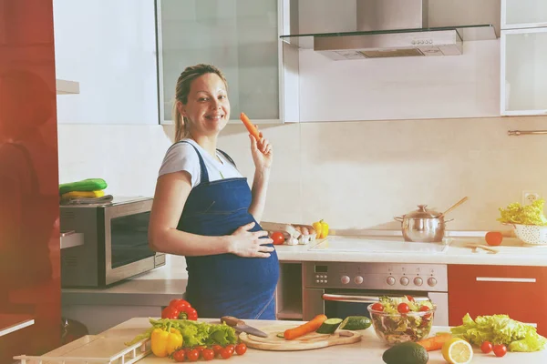 Těhotná žena jíst mrkev v kuchyni — Stock fotografie