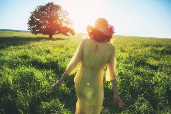 美しいです黄色のドレスと帽子夏のフィールドで日光の下で歩く女性 — ストック写真