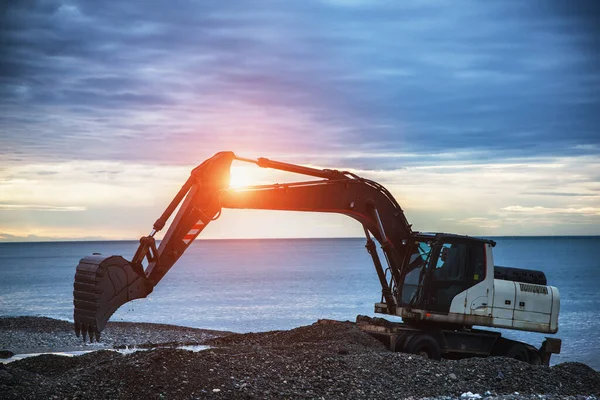 日の出の光の中で海の近くの工業地帯の掘削現場でバケツと働くバックホーや掘り人 — ストック写真