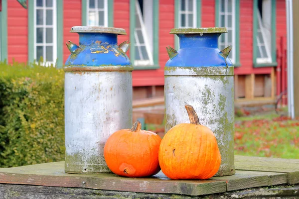 Zwei Historische Milchkannen Und Orangefarbene Kürbisse Vor Roten Bauernhäusern — Stockfoto