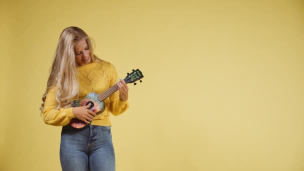 Vista frontal de una mujer rubia tocando un Ukulele con un fondo amarillo — Vídeo de stock