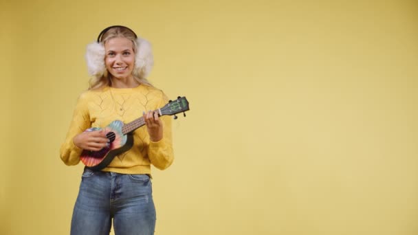 Mujer joven artística tocando el Ukulele mientras usa auriculares esponjosos — Vídeos de Stock