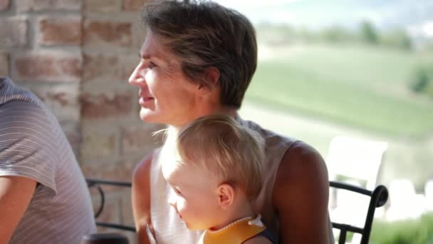 Стара леді тримає дитину розмовляючи з кимось під час сімейної вечері — стокове відео