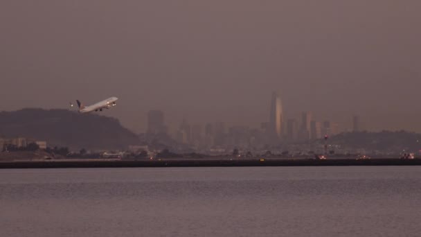 Avião decolando durante o pôr do sol com luzes da cidade em segundo plano — Vídeo de Stock