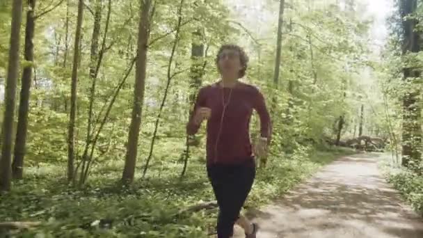 Aktywna kobieta biegająca po lesie wzdłuż ścieżki nosząc słuchawki — Wideo stockowe