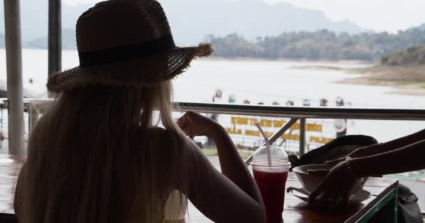 Женщина в одиночестве пьет коктейль и смотрит на озеро — стоковое видео