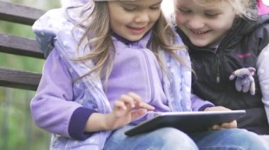 Tablet bilgisayar ile iki küçük kız bahçede bankta oturur