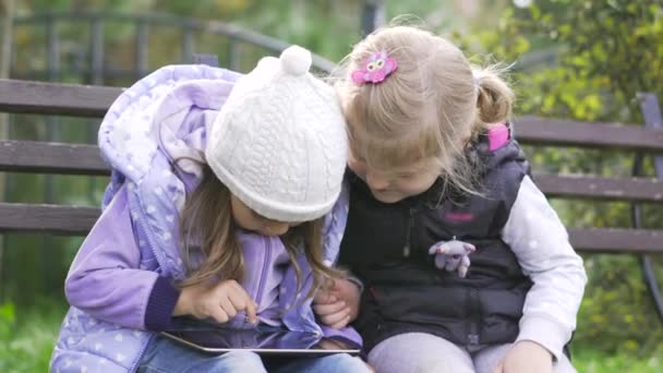 Две маленькие девочки с планшетным компьютером сидят на скамейке в саду — стоковое видео