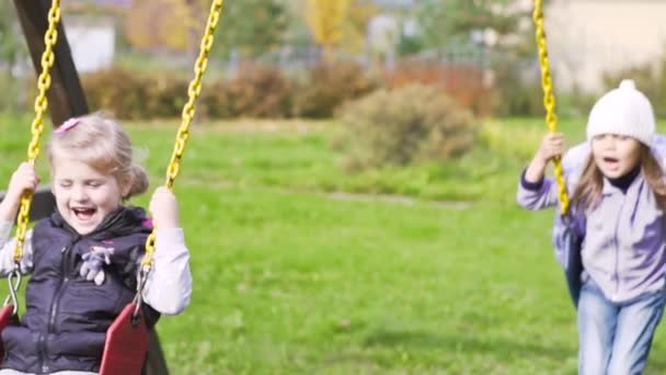 Zwei schöne kleine Mädchen auf einer Schaukel im Freien auf dem Spielplatz — Stockvideo