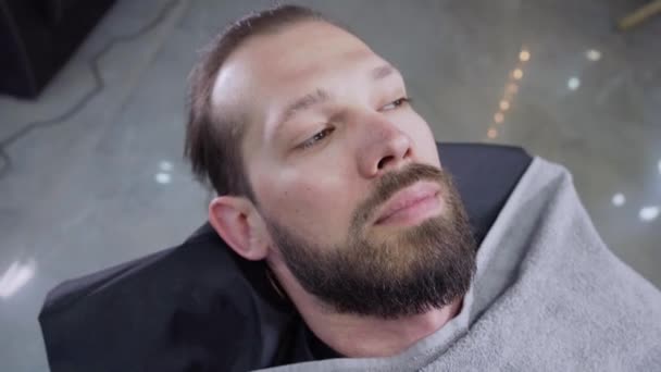 Happy young man with a beard lies in a barbers chair and smiling. — Stock Video