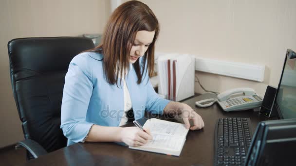 Businesswoman brunette in a blue jacket smiles and writes in her diary in the office. — Stock Video